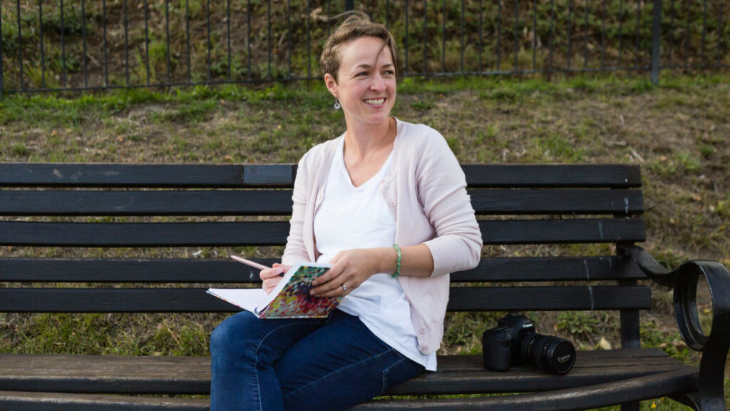 Woman writing in notebook on park bench with camera.