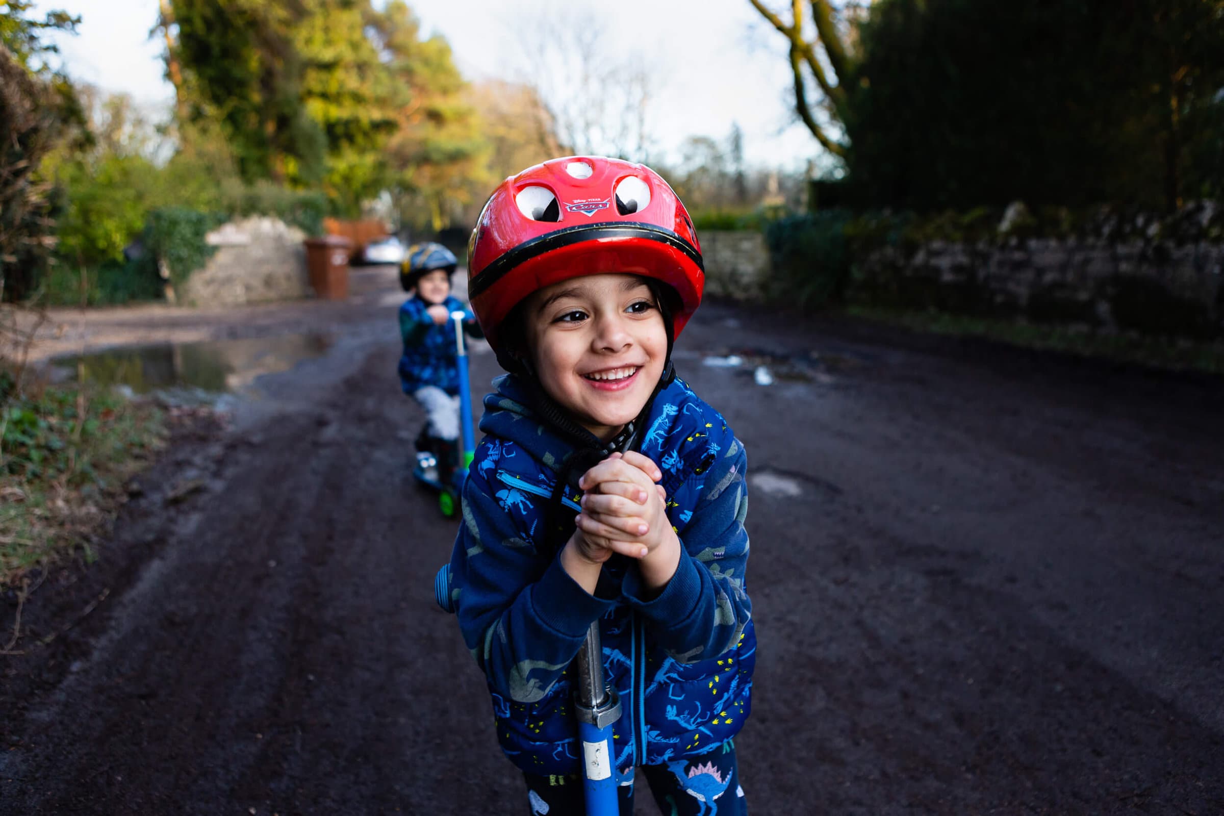 A very happy looking young boy on his scooter with his brother behind | Family photographer Bristol