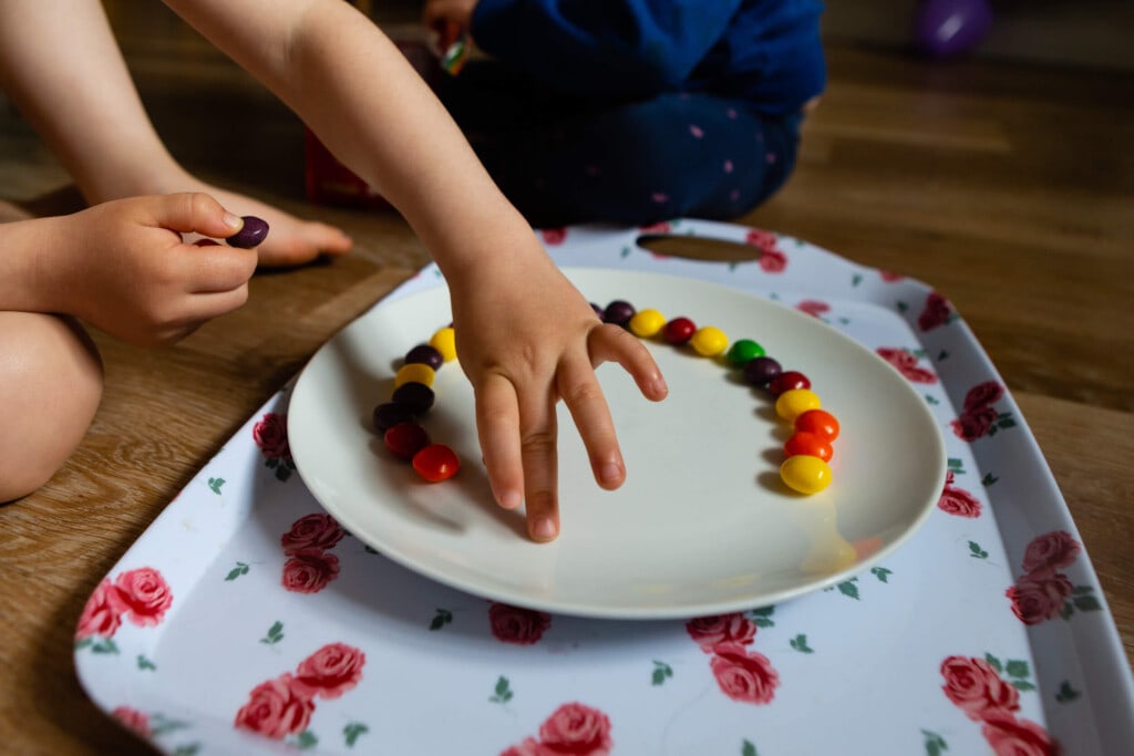 A child’s hand arranges colorful candies in a circle on a white plate placed on a floral tray, with another child sitting cross-legged in the background on a wooden floor. Rose Dedman Photography