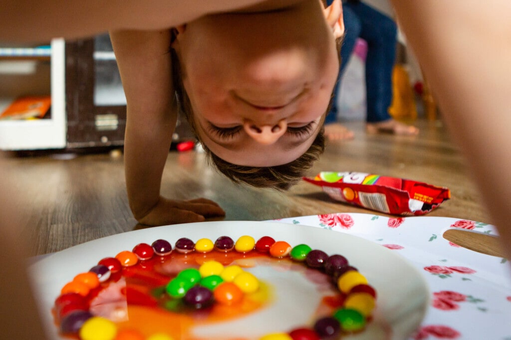 A child leans over a plate of colorful candy arranged in a circle, with candies dissolving in water to create a vibrant rainbow pattern. The scene is playful and set indoors on a wooden floor. Rose Dedman Photography
