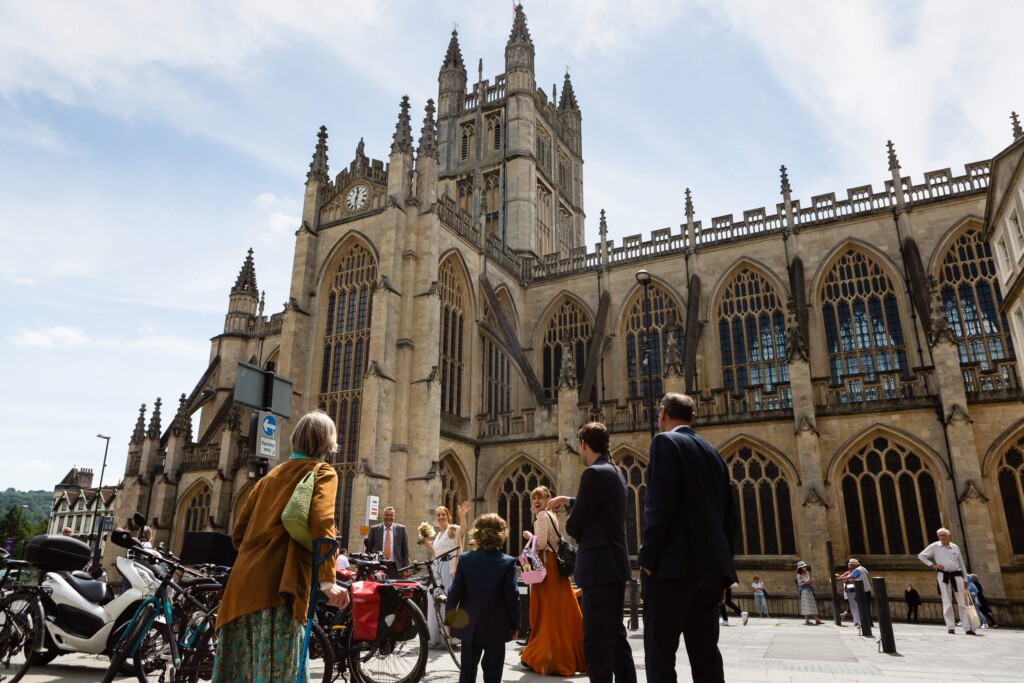 A group of people in formal attire stands outside Bath Abbey, a large Gothic church with tall spires and arched windows, on a sunny day. Bicycles are parked nearby. Rose Dedman Photography