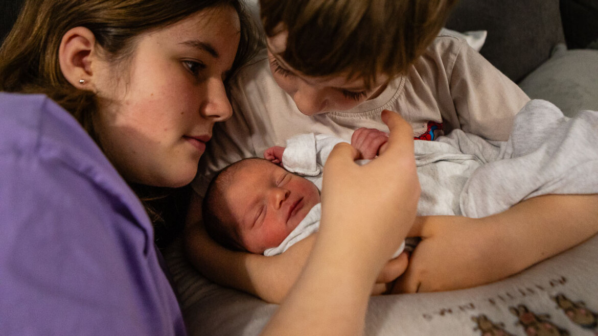 Two children gently cuddle a newborn baby, who is wrapped in a white blanket and sleeping peacefully in their arms. The scene is warm and intimate, showing a close bond between the siblings. Rose Dedman Photography