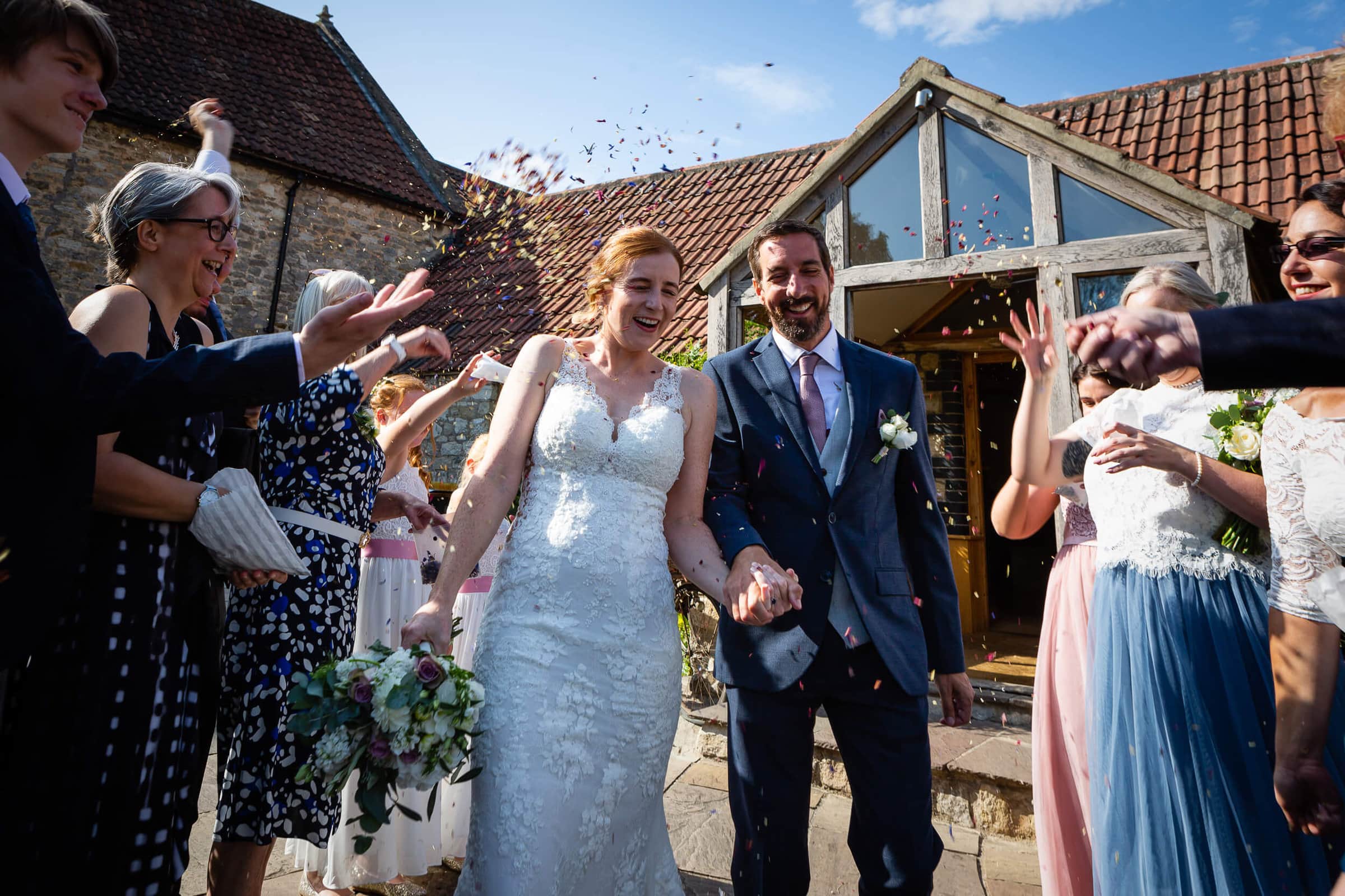 A joyful bride and groom walk hand in hand outside, smiling as guests throw confetti. The bride wears a white lace gown and holds a bouquet; the groom is in a suit. Friends and family celebrate under a sunny sky. Rose Dedman Photography