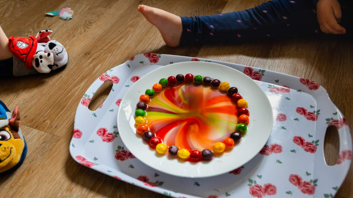 A plate with colorful candies arranged in a circle sits on a floral tray; the candies’ colors are swirling in the center. Two children’s legs and stuffed toys are visible nearby on a wooden floor. Rose Dedman Photography