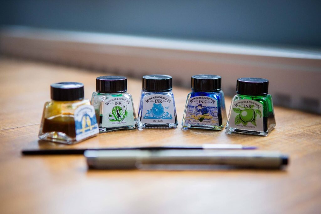 Five small bottles of Winsor & Newton ink in various colors are lined up on a wooden surface with a paintbrush and pen in front—an inviting scene perfect for commercial photography. The softly blurred background adds artistic flair. Rose Dedman Photography