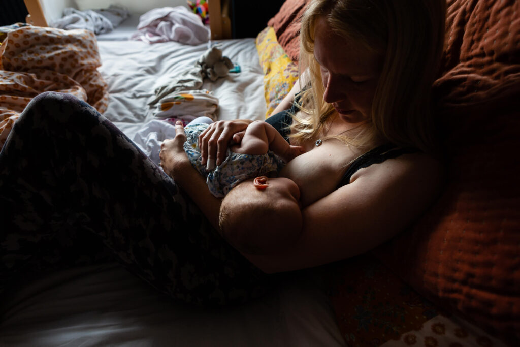 A woman sits on a bed, breastfeeding her baby in a softly lit room. The calm, intimate atmosphere is reminiscent of the gentle style found in natural wedding photography—full of warmth and loving connection. Rose Dedman Photography
