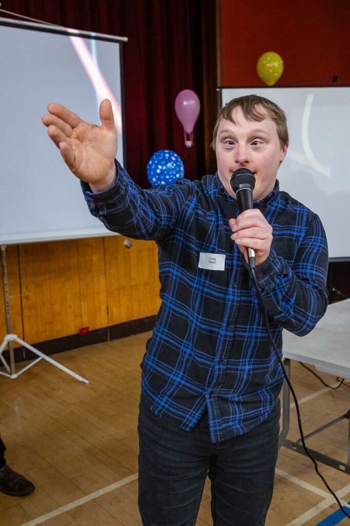 A man with Down syndrome wearing a blue plaid shirt holds a microphone and gestures while speaking at an indoor event. Colorful balloons and projection screens in the background highlight the engaging charity photography setting. Rose Dedman Photography