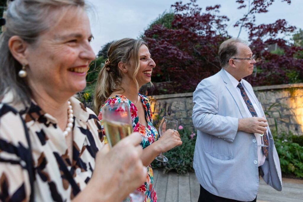 Three adults stand outdoors, smiling and holding champagne glasses. Perfect for event photography, two women in patterned dresses are in the foreground, while a man in a light jacket and tie stands beside them. Trees and a stone wall are behind them. Rose Dedman Photography