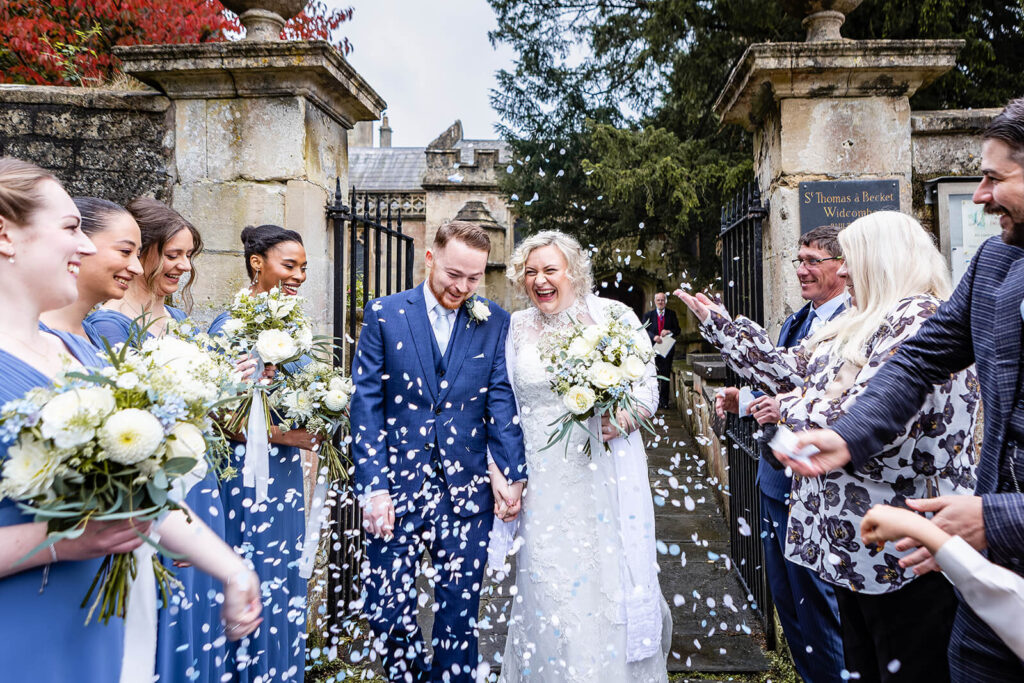 A bride and groom walk hand in hand outside, smiling as guests throw white confetti after a beautiful Church Wedding Bath. Bridesmaids in blue dresses hold bouquets, and everyone looks joyful at the celebration outside a historic stone church. Rose Dedman Photography
