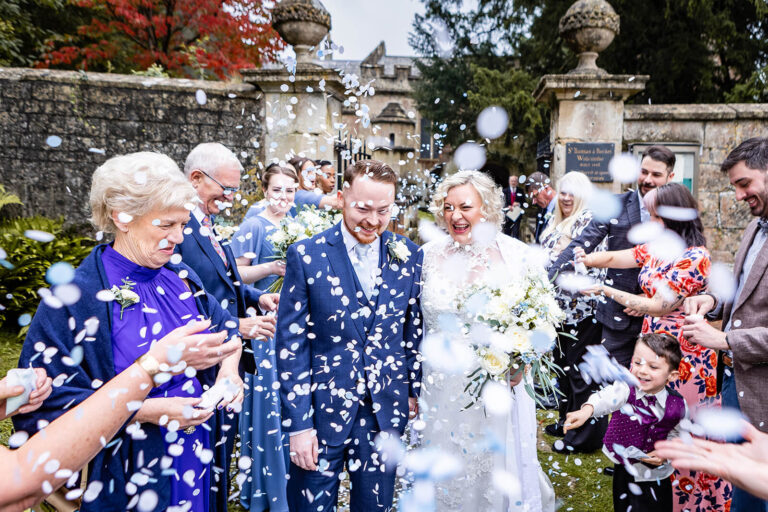 A bride and groom walk outside among guests who joyfully throw white confetti. The Bath wedding couple smiles brightly, surrounded by family and friends, with greenery and a stone building in the background. Rose Dedman Photography