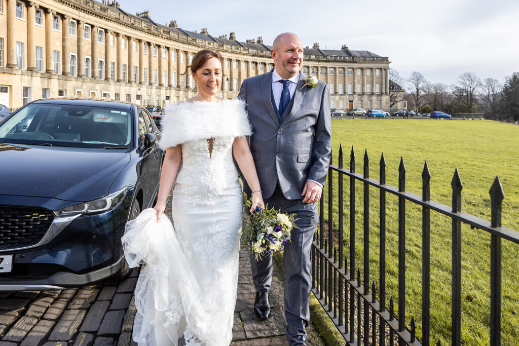 A bride in a white dress and groom in a gray suit walk hand in hand outdoors, smiling, with a bouquet and historic curved buildings—perfect for Guildhall Bath wedding photos. A parked car and iron fence are nearby on a sunny day. Rose Dedman Photography