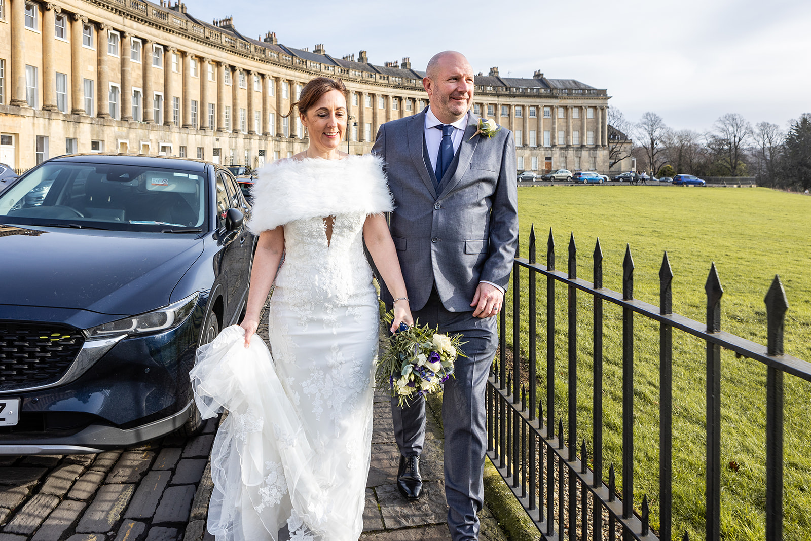 A bride in a white dress and groom in a gray suit walk hand in hand outdoors, smiling, with a bouquet in front of Bath's Royal Crescent. Somerset Wedding Photographer Rose Dedman