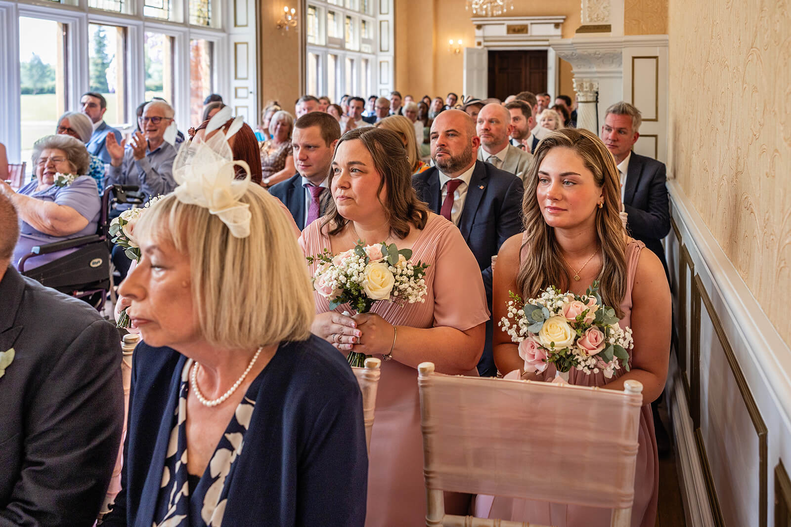 Wedding guests seated with flower bouquets at Holmewood Hall wedding ceremony. Wedding photographer Rose Dedman.