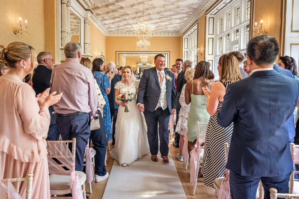 Bride and groom walking down the aisle, smiling after the ceremony at Holmewood Hall country house wedding venue. 