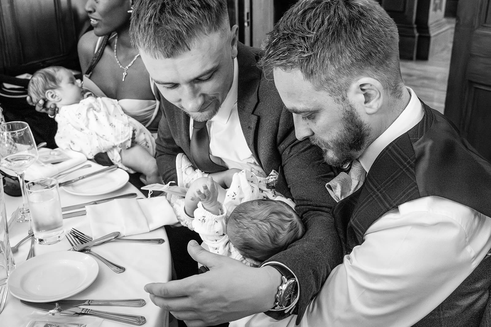Two men hold babies at a table at Holmewood Hall wedding. 