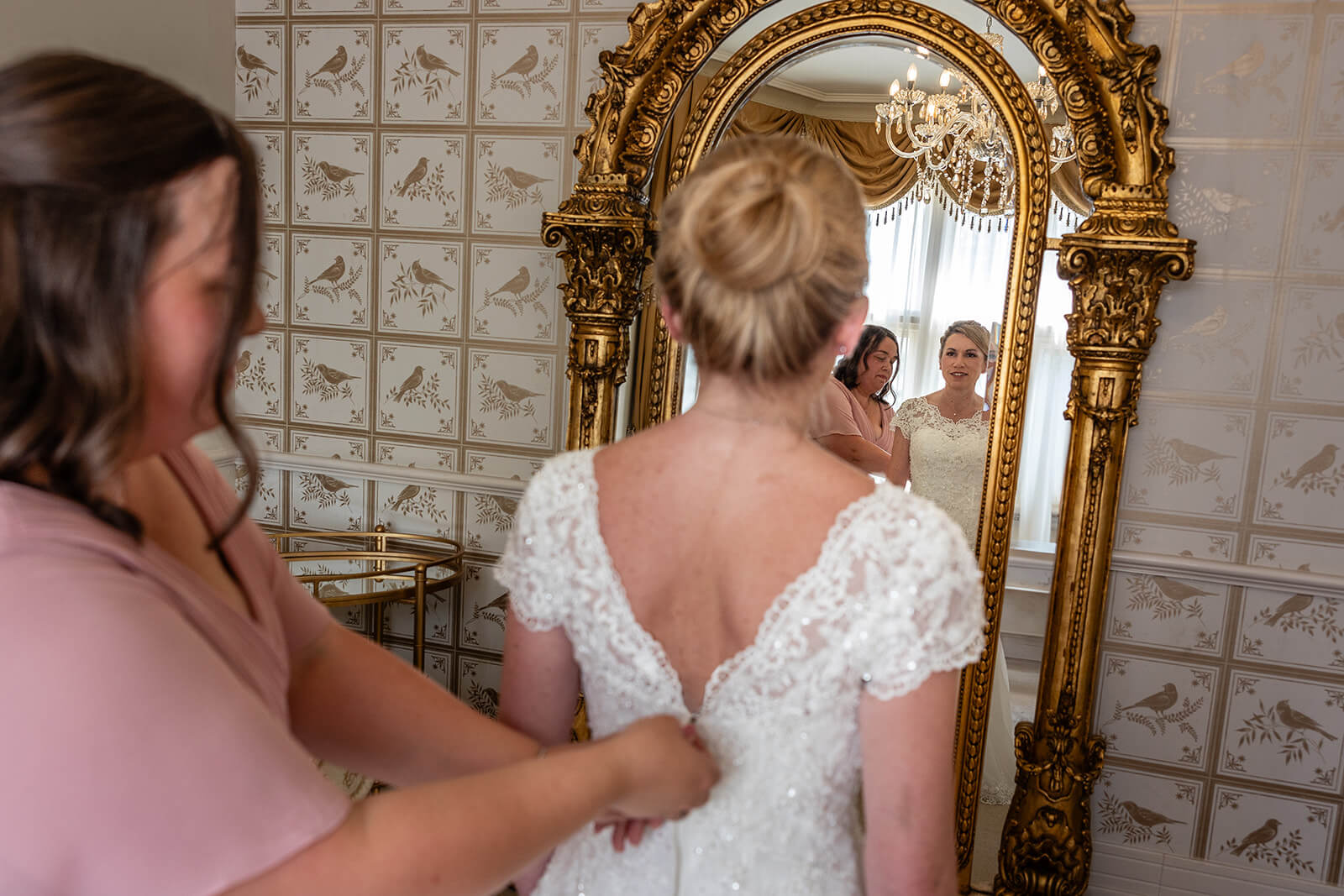 Bride getting dressed reflected in ornate mirror. Country house wedding at Holmewood Hall.