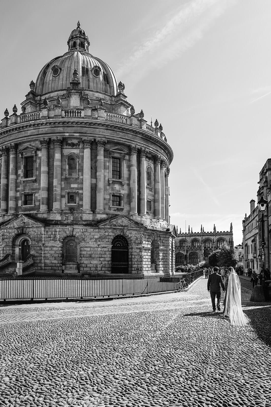The Radcliffe Camera in Oxford with bride and groom walking to the side, in black and white | Oxford Nikkah wedding photos